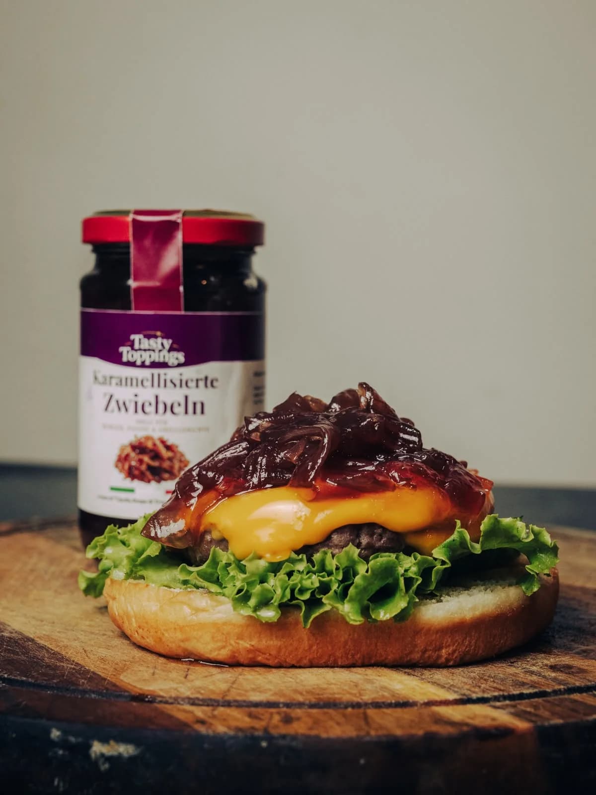 Offener Burger mit Salat, geschmolzenem Käse und karamellisierten Tropea-Zwiebeln auf einem Holzbrett, dahinter das Glas Tasty Toppings